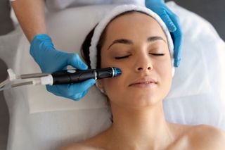 Woman receiving a facial treatment with a handheld device. Gloved hands, white headband, eyes closed.