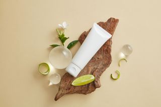 White skincare tube on wood, lime slice, flowers, and glass spheres on a beige background.