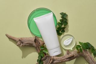 White cosmetic tube, sample of cream, green leaves on driftwood against a light green background.
