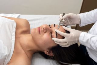 Woman getting eyebrow shaping with tweezers on a white sheet, by gloved hands.