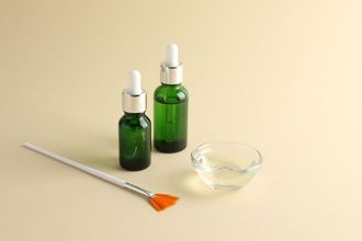 Two green serum bottles with droppers, brush, and clear liquid in bowl, on beige surface.