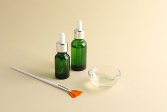 Two green serum bottles with droppers, a brush, and a bowl of liquid on a beige surface.