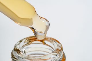 Dropper dispensing golden liquid into a clear glass bottle, close up on a white background.