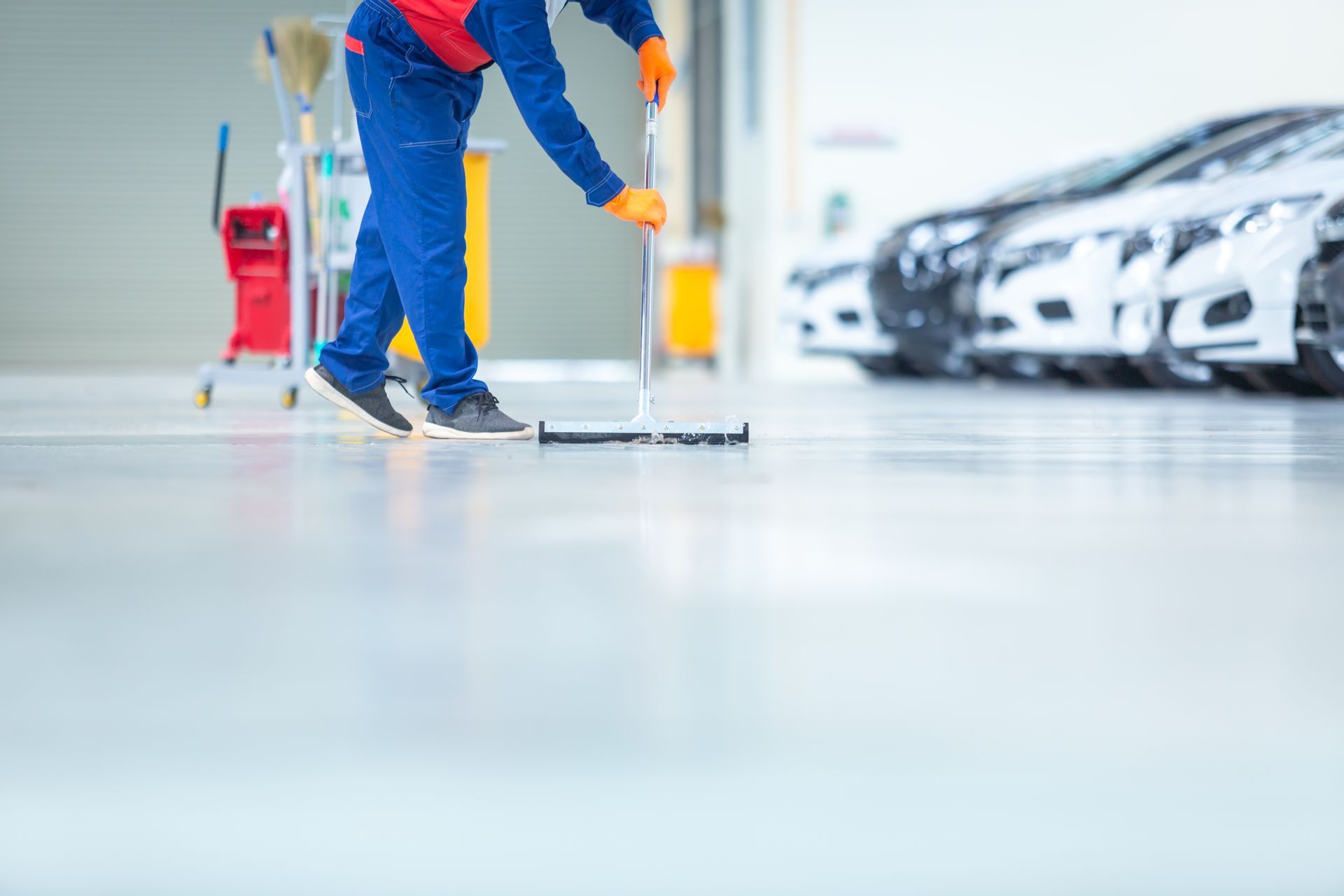 Person sweeping a shiny gray floor in a car dealership. Cars are parked in the background.