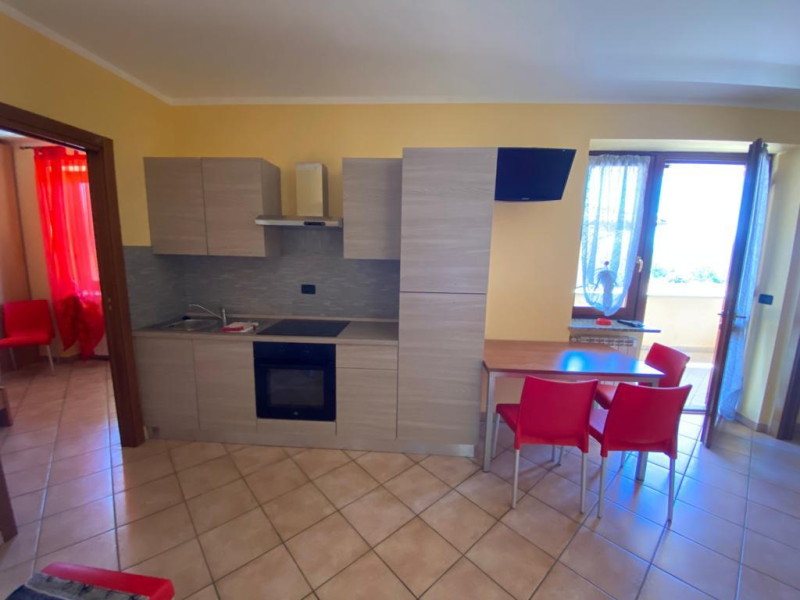 Cucina con mobili beige, sedie rosse e porta aperta su un balcone.