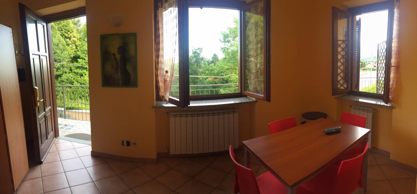 Stanza interna con porta e finestre aperte. Tavolo con sedie. Pareti gialle e luminose.