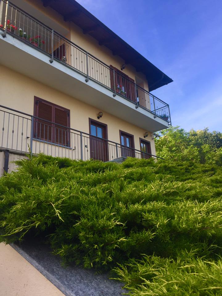 Casa beige con balcone e persiane marroni, parzialmente nascosta da cespugli verdi, contro un cielo azzurro.