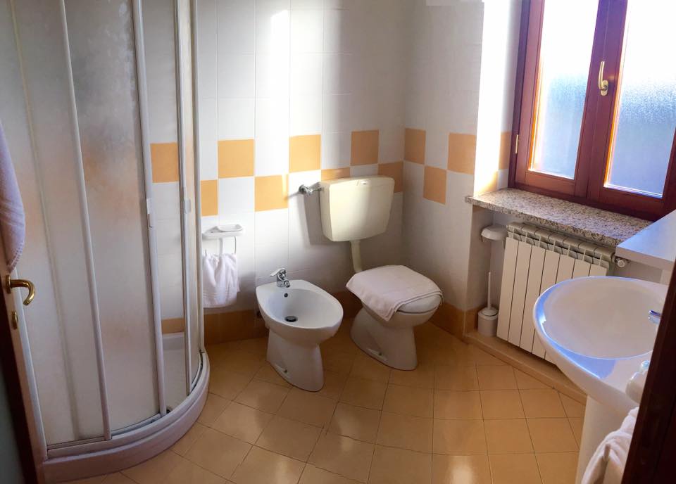 Bagno con doccia, WC, bidet e lavandino, in una stanza con finestra. Piastrelle a scacchi gialle e bianche.