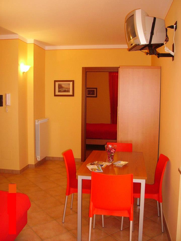 Zona pranzo con pareti arancioni, tavolo, sedie, TV e porta aperta su una stanza rossa.