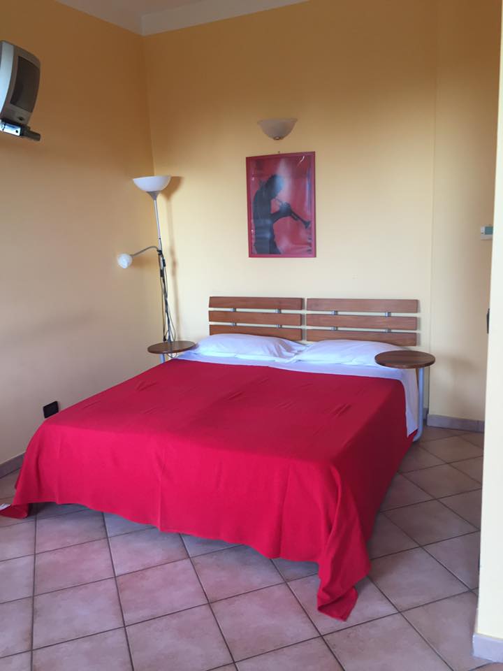 Camera da letto con copriletto rosso, testiera in legno, comodini, lampada da terra e opere d'arte sulla parete gialla.