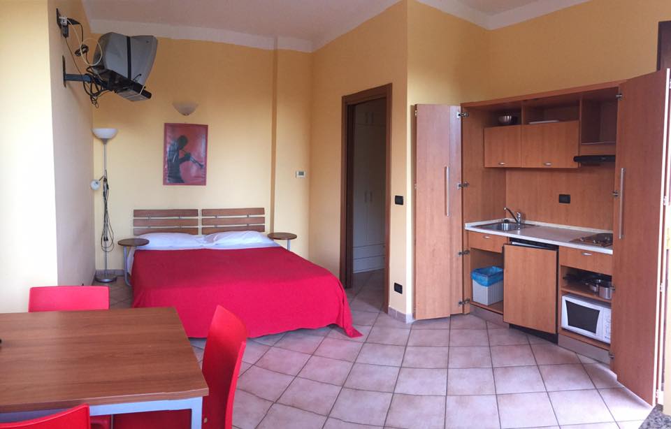 Piccolo monolocale giallo con letto rosso, cucina compatta e tavolo.