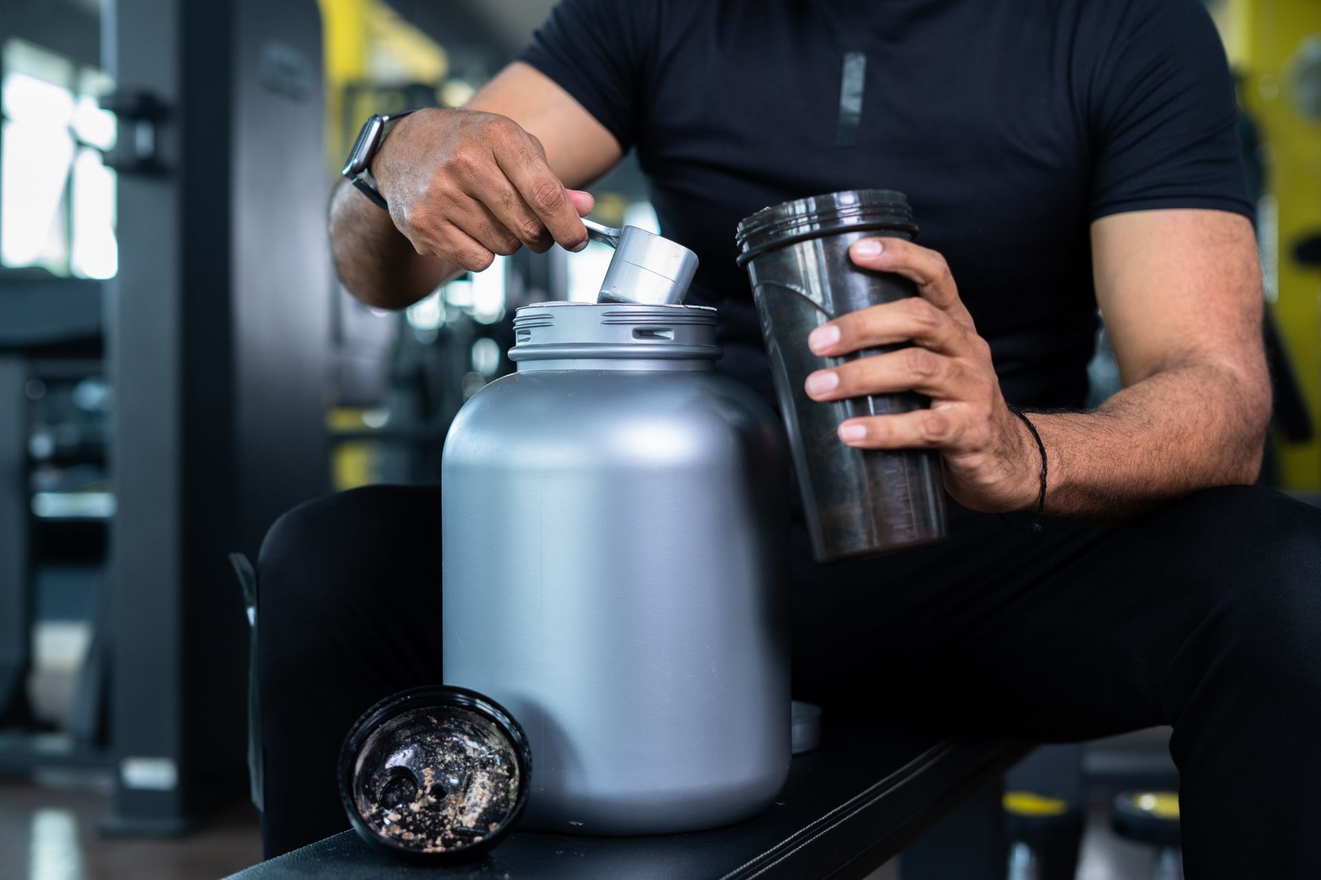 A man is sitting on a bench in a gym preparing a protein shake.