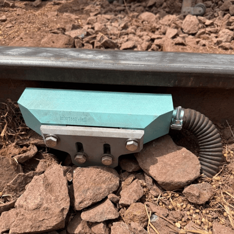 A teal-colored railway rail lubricator attached to a track, surrounded by rocks and dirt.