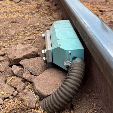 A teal-colored railway component with a corrugated tube attached to a rail on a gravel bed.