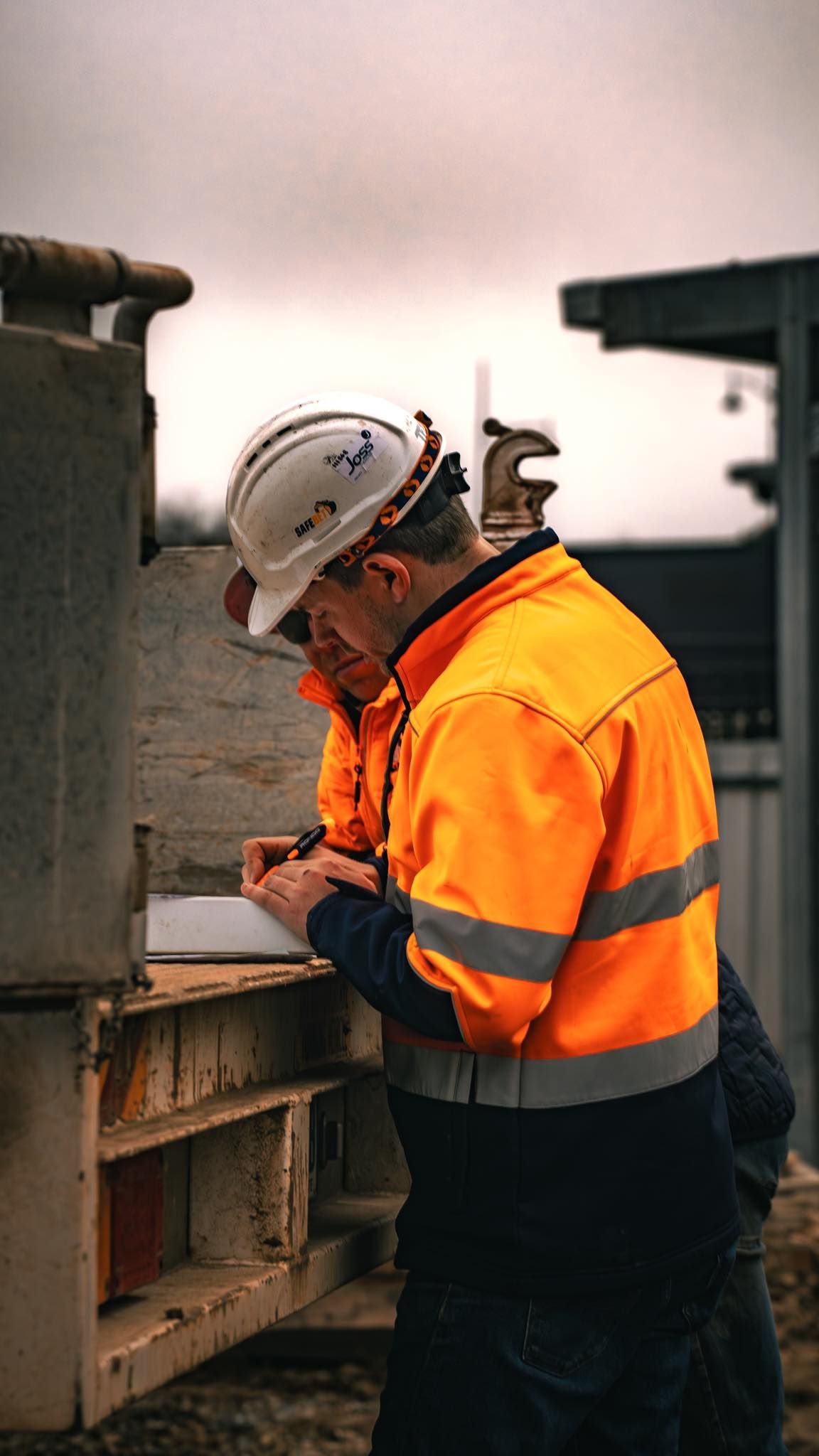 Construction worker in orange safety jacket and hard hat, looking at paperwork at a construction site.