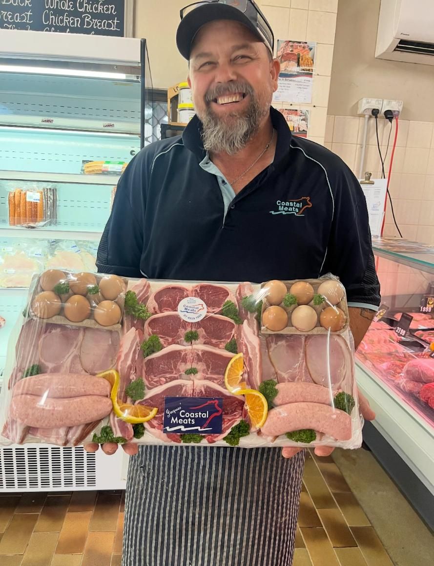 Butcher in a Black Shirt and Apron Smiles, Holding a Packed Meat Platter in a Butcher Shop — Coastal Meats in Yeppoon, QLD