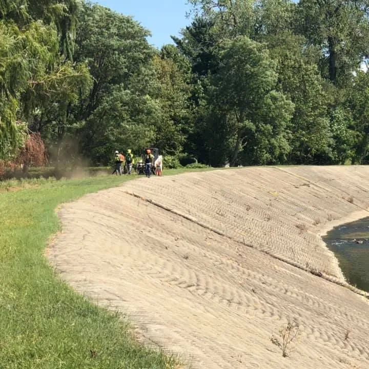 A group of people are standing on a dirt hill next to a body of water.
