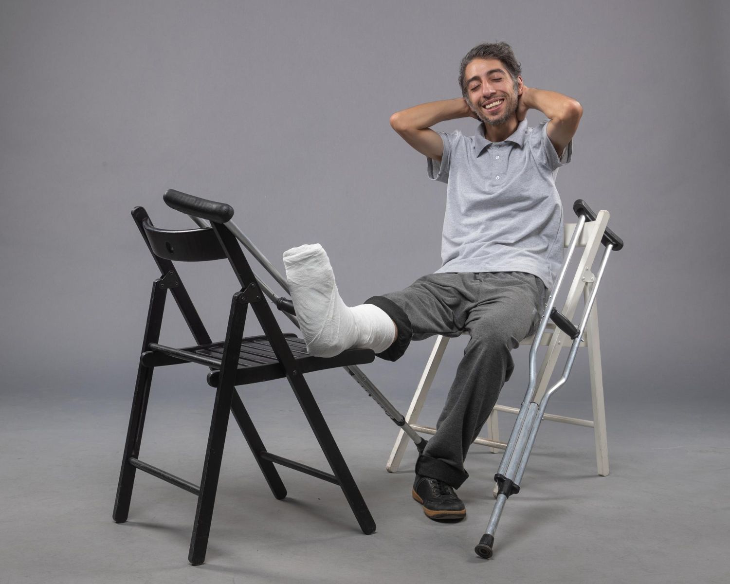 Man with leg in cast rests on chair, smiling, crutches nearby. Gray background.