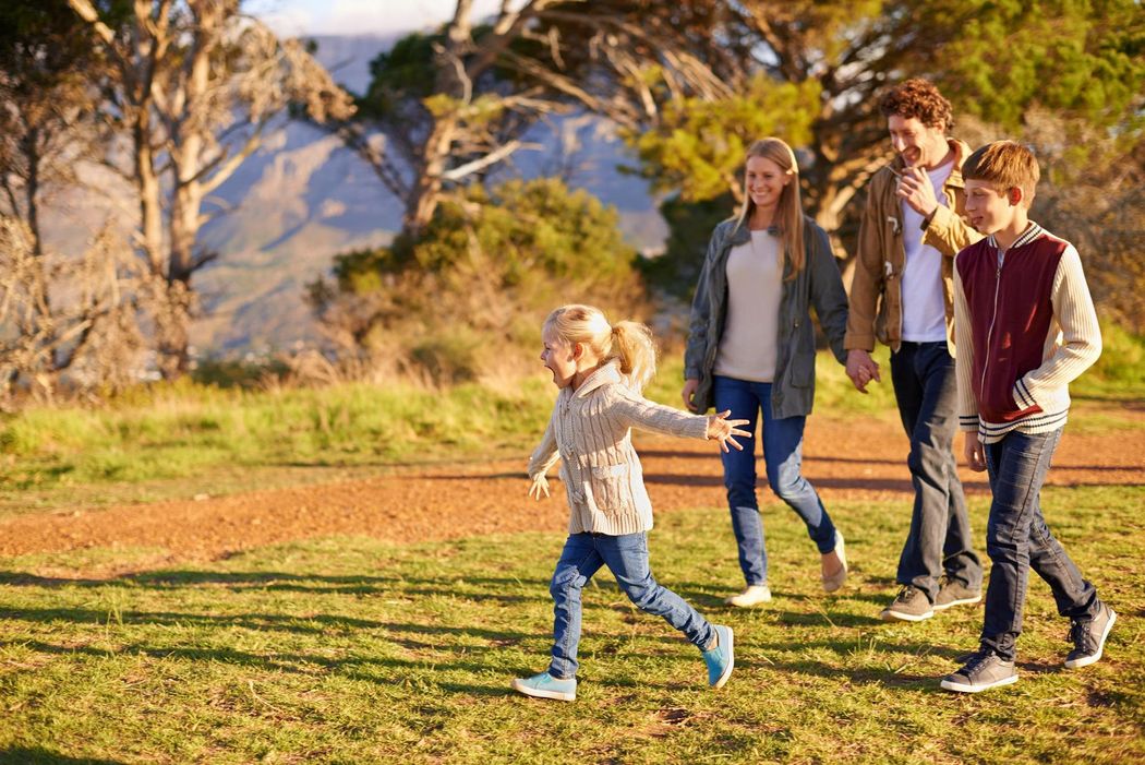 Family hiking, child running ahead on a grassy path with mountains and trees in the background.