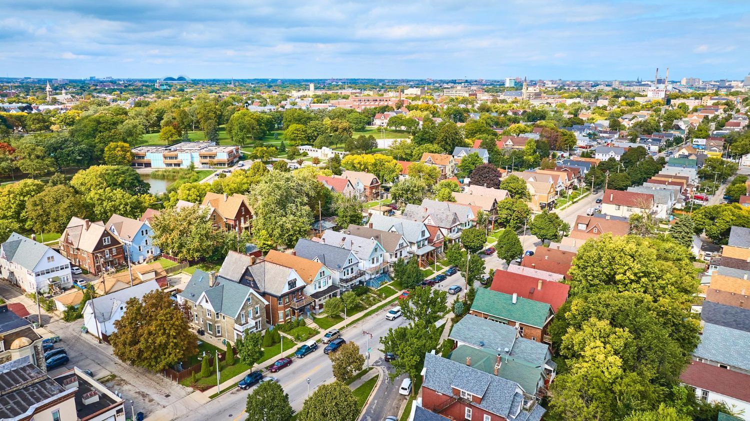Aerial view of a neighborhood with colorful houses, mature trees, and a street. Sunny, blue sky.
