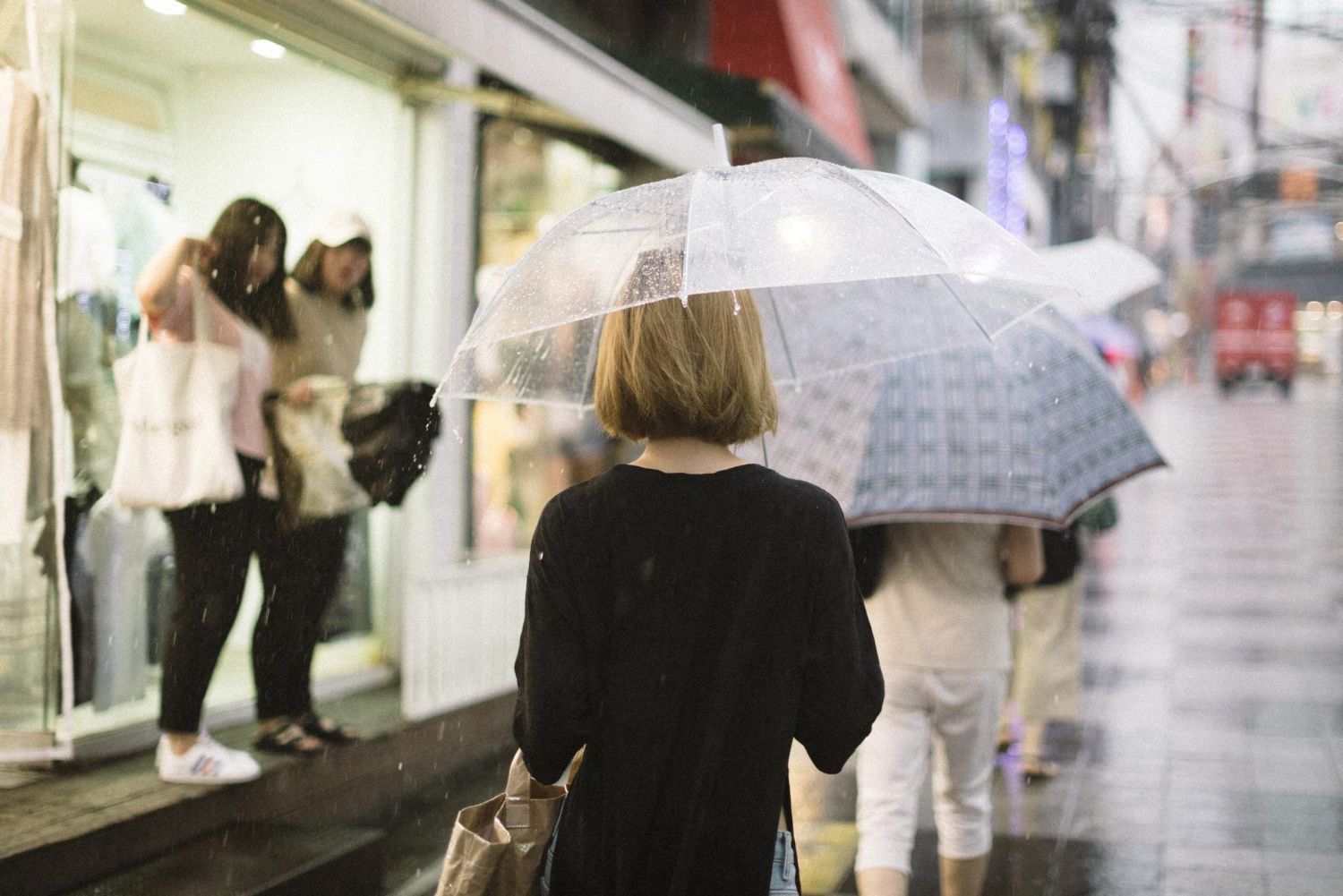 Person with umbrella walks on a rainy street past a storefront with other people visible.
