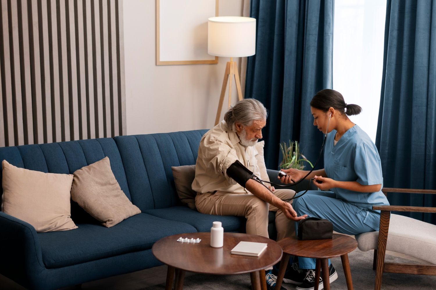 A healthcare worker checks a patient's blood pressure on a sofa in a living room.