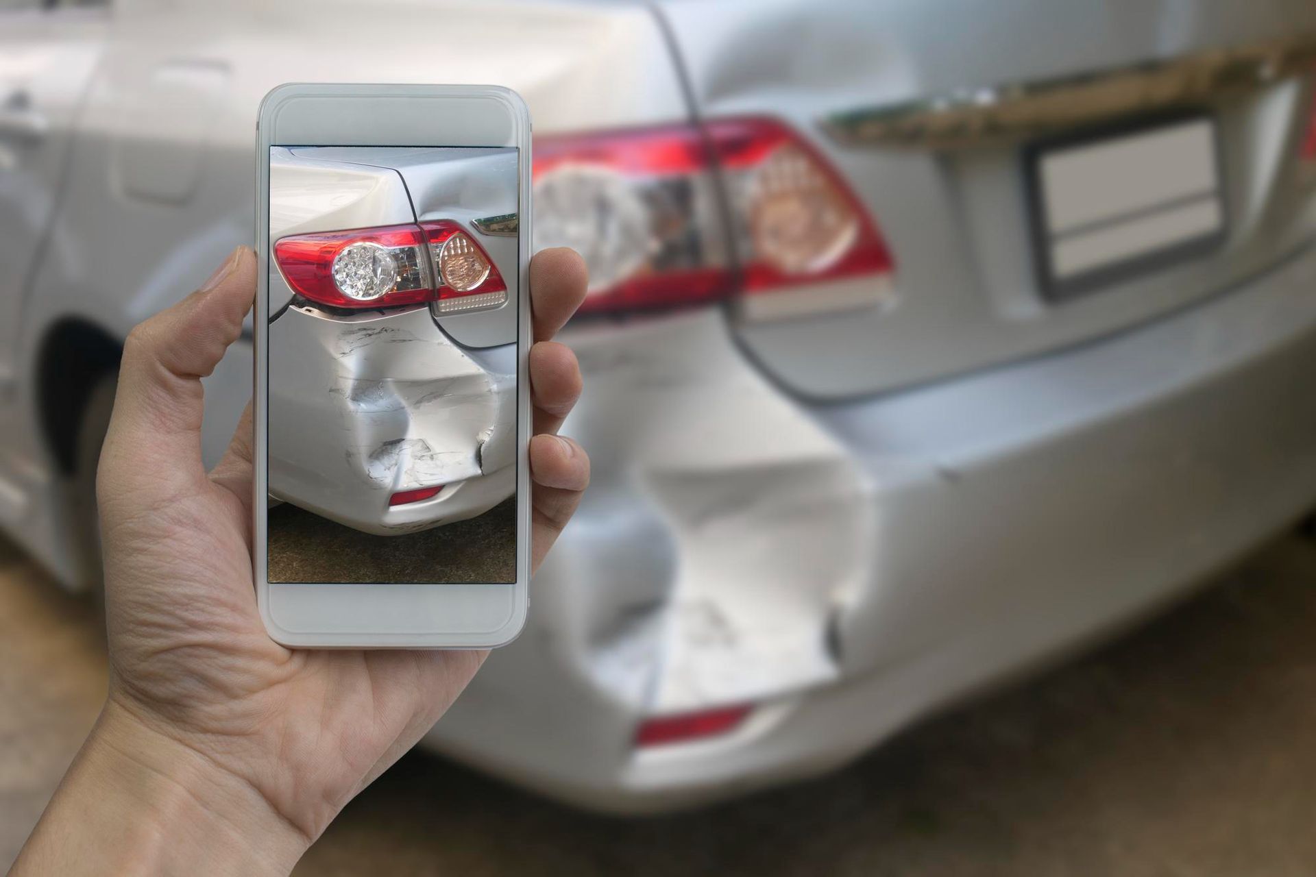 Hand holding phone displaying a photo of a silver car with rear-end damage.