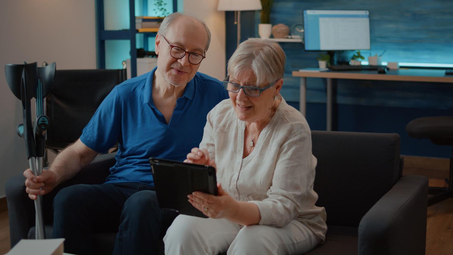 An elderly couple on a couch looking at a tablet together, one holding a crutch.