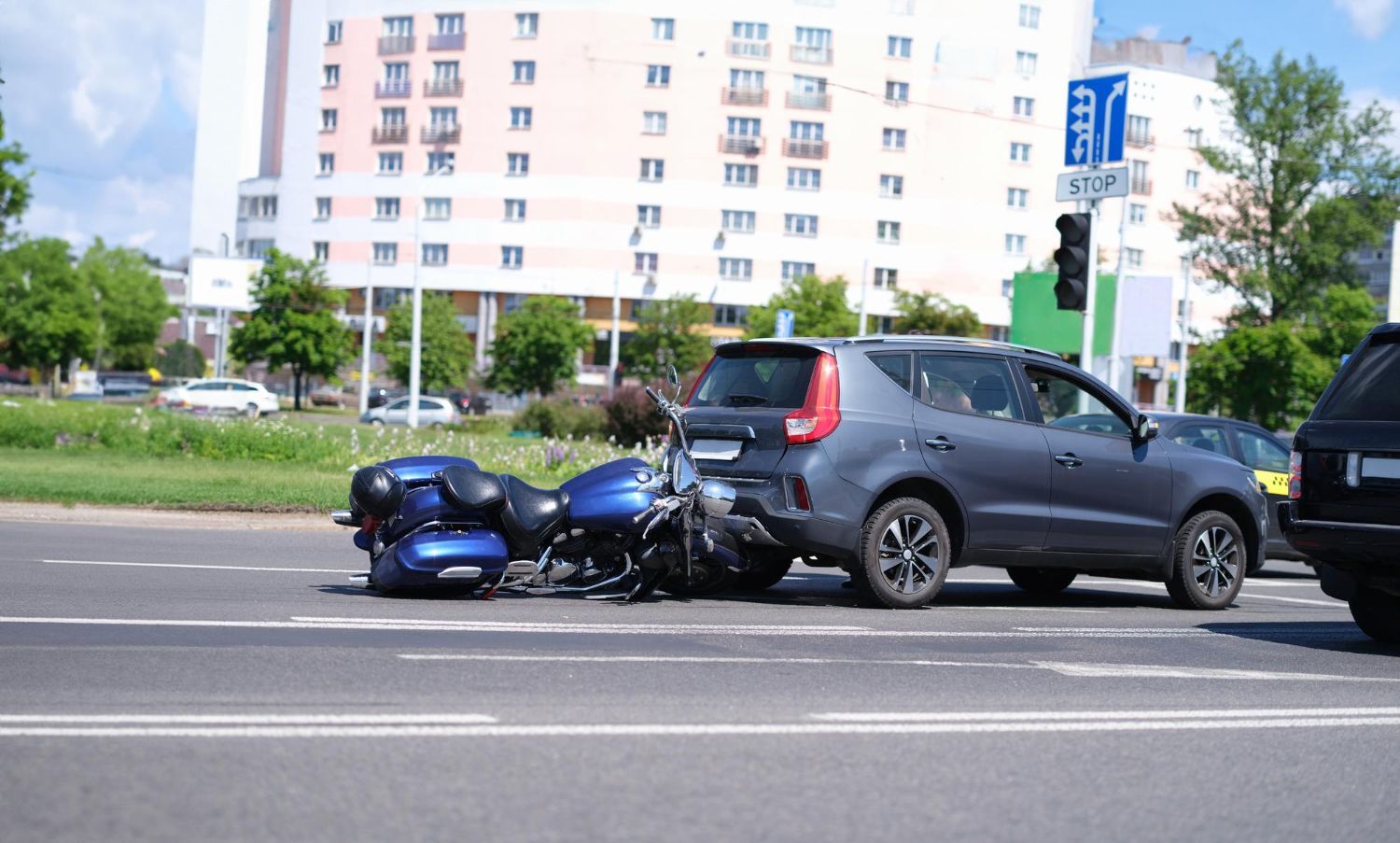 Motorcycle and car accident on a city street; blue motorcycle damaged, gray SUV with rear damage.