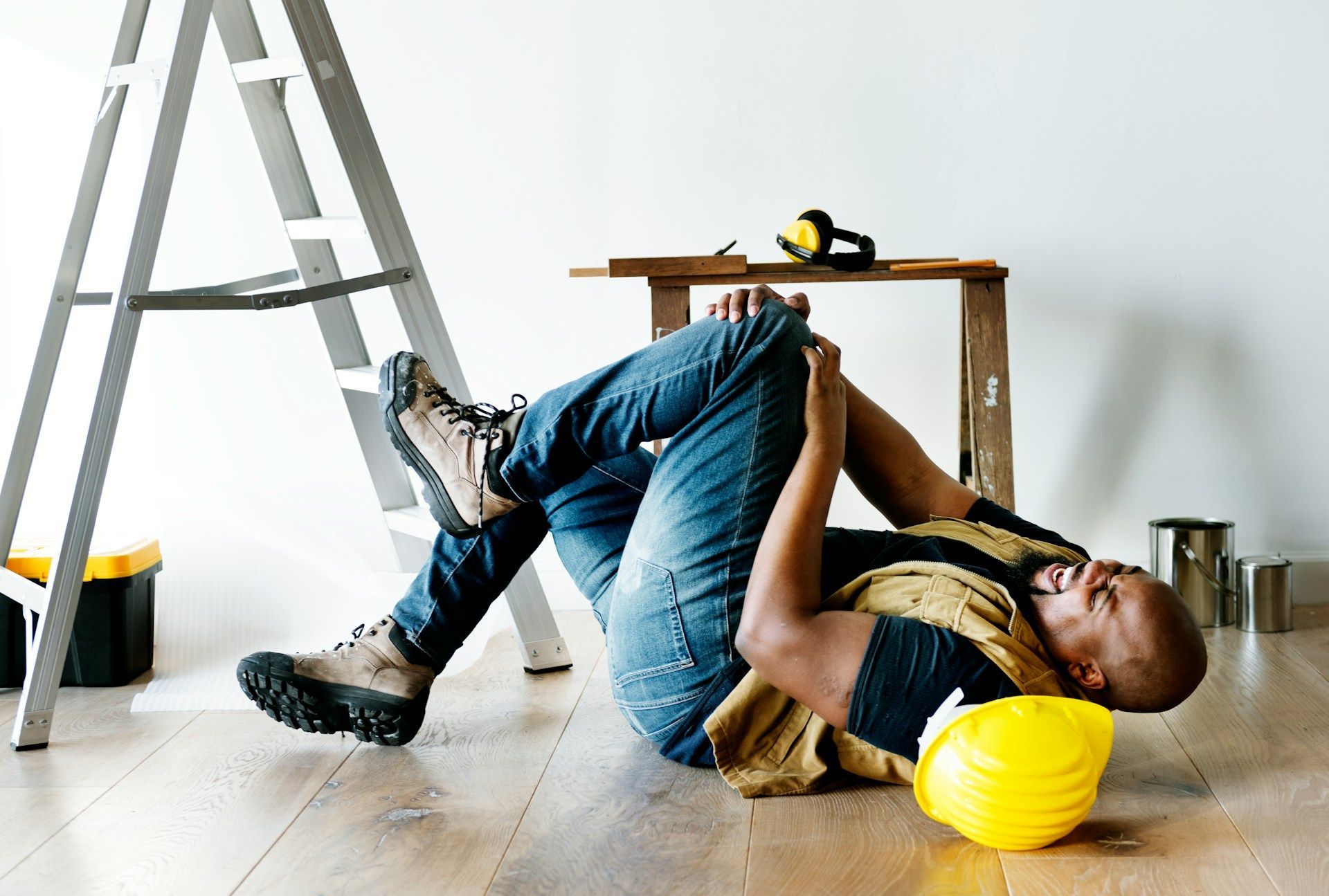 Man lying on floor, grimacing, near a ladder and yellow hard hat.