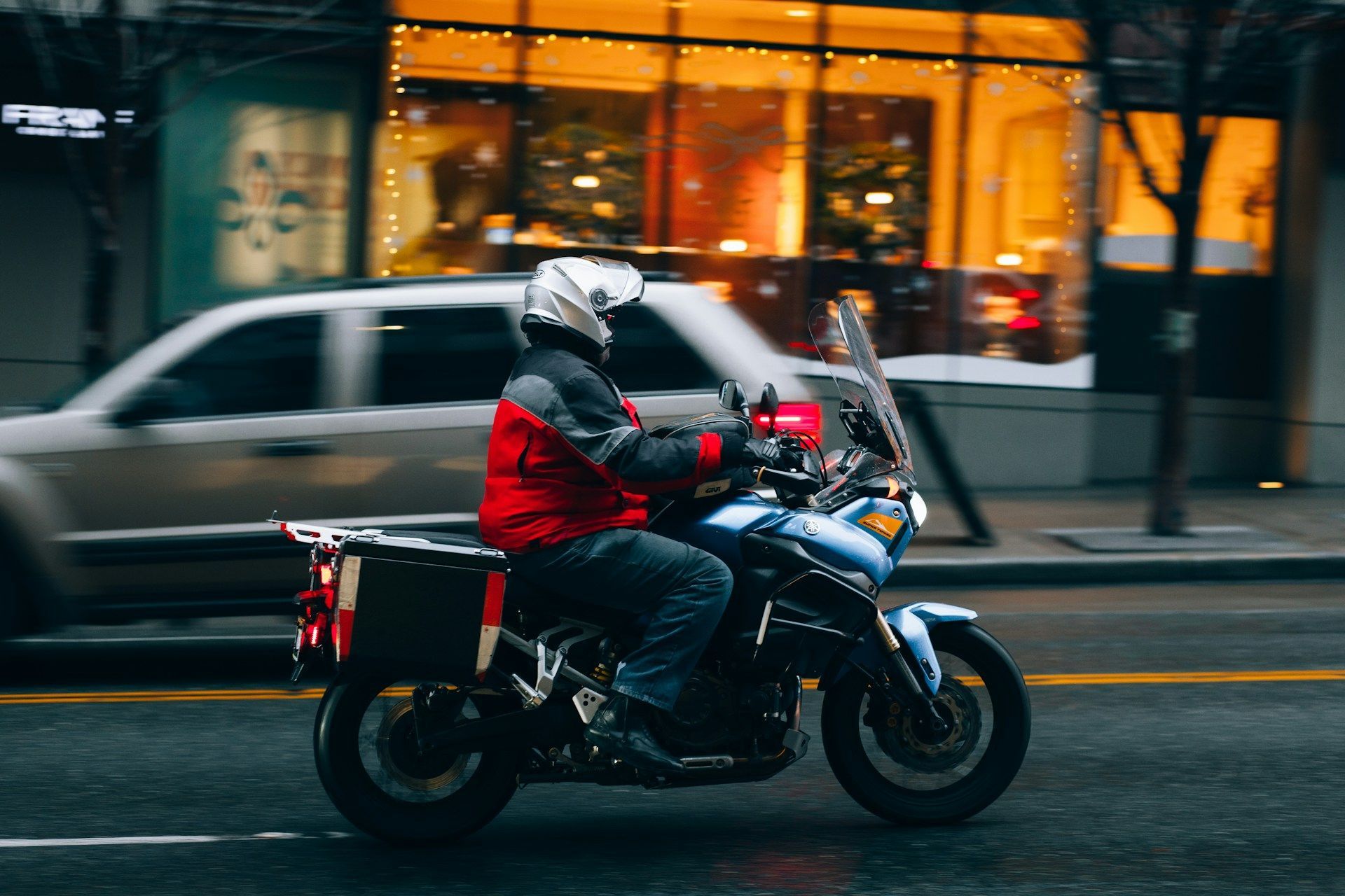 Motorcyclist in red jacket and helmet riding blue motorcycle on city street, blurred background.
