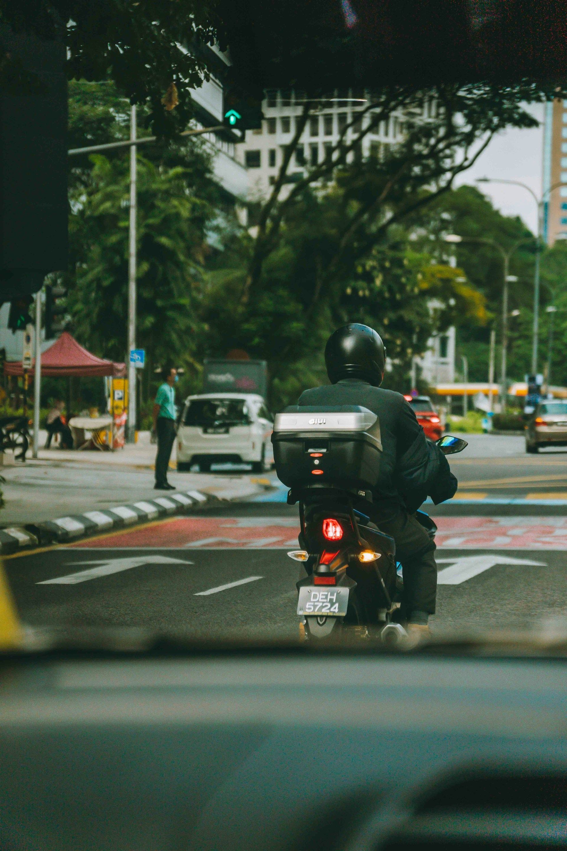 Motorcyclist in black jacket with a cargo box on city street with traffic and a building.