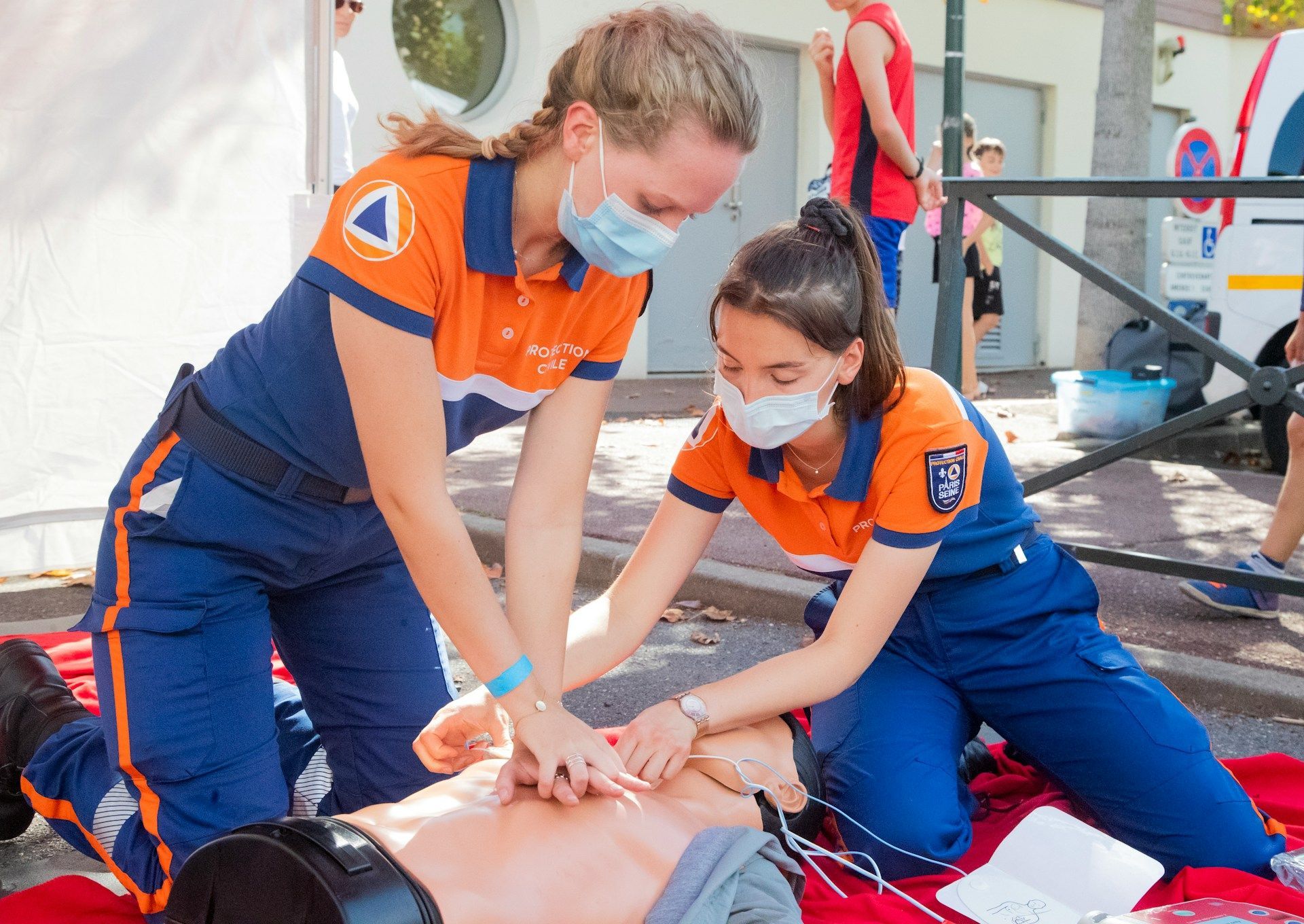 Two people in orange/blue uniforms demonstrating CPR on a mannequin outdoors.