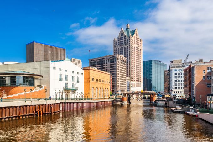 Downtown Milwaukee skyline along a river; blue sky, buildings of various heights and materials, water reflection.