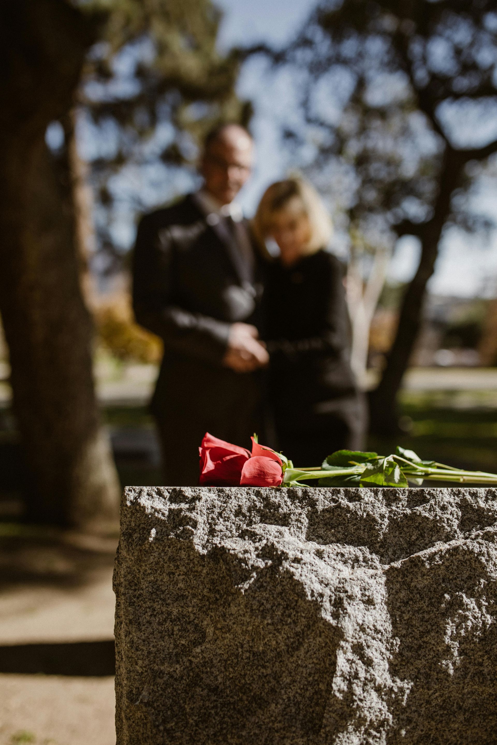 A red rose on a gravestone with two people mourning in the background at a cemetery.