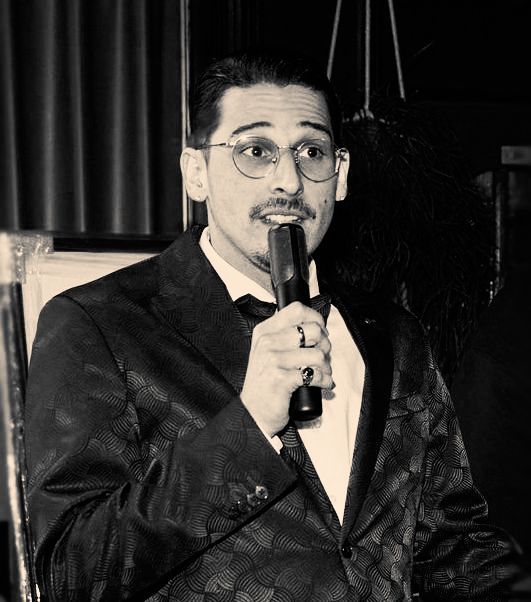 Een man in formele kleding, met een bril op, spreekt in een microfoon in een schemerig verlichte binnenruimte.