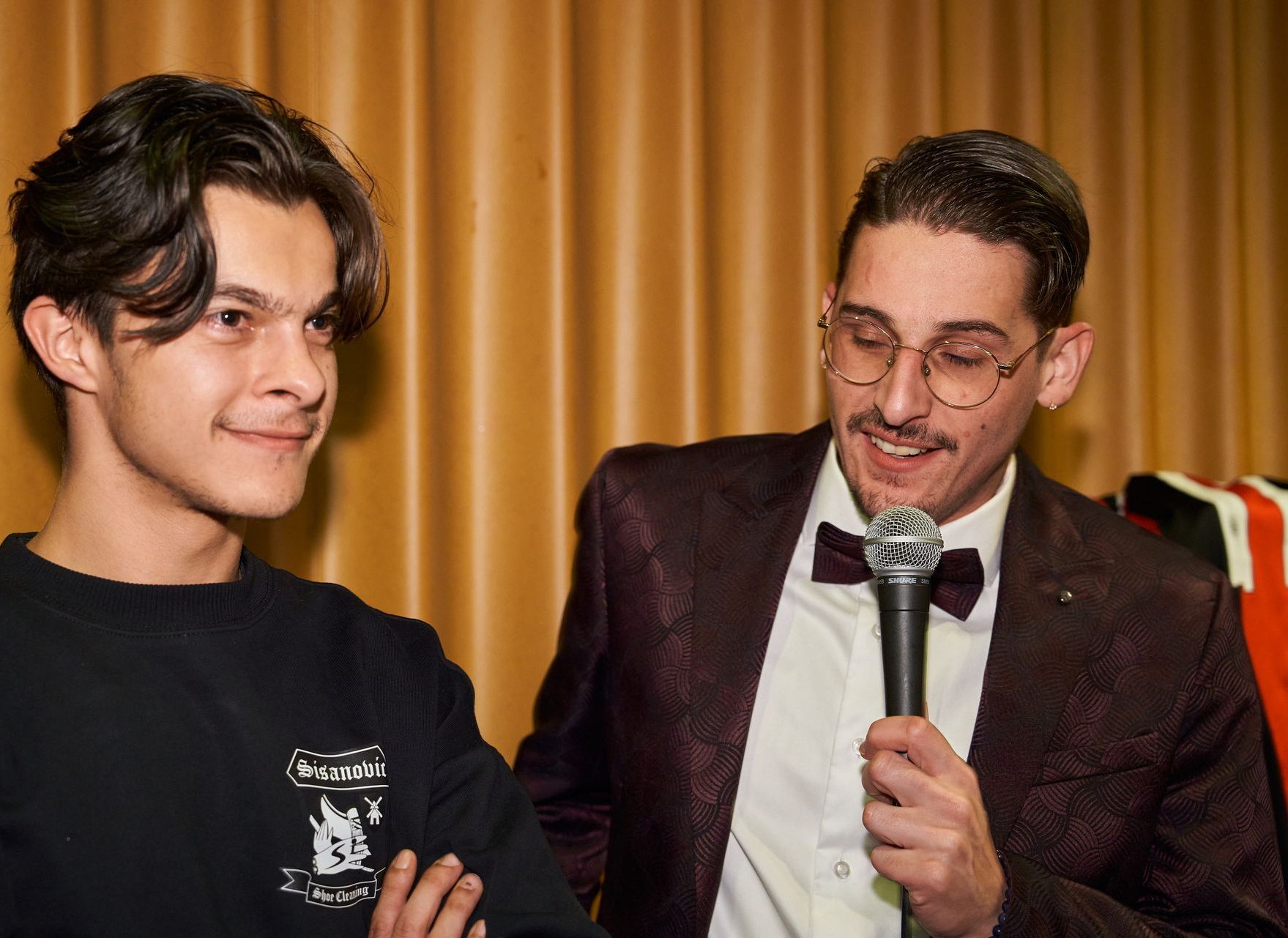 Twee mannen, van wie er &eacute;&eacute;n een microfoon vasthoudt, staan ​​lachend bij een toneelgordijn.