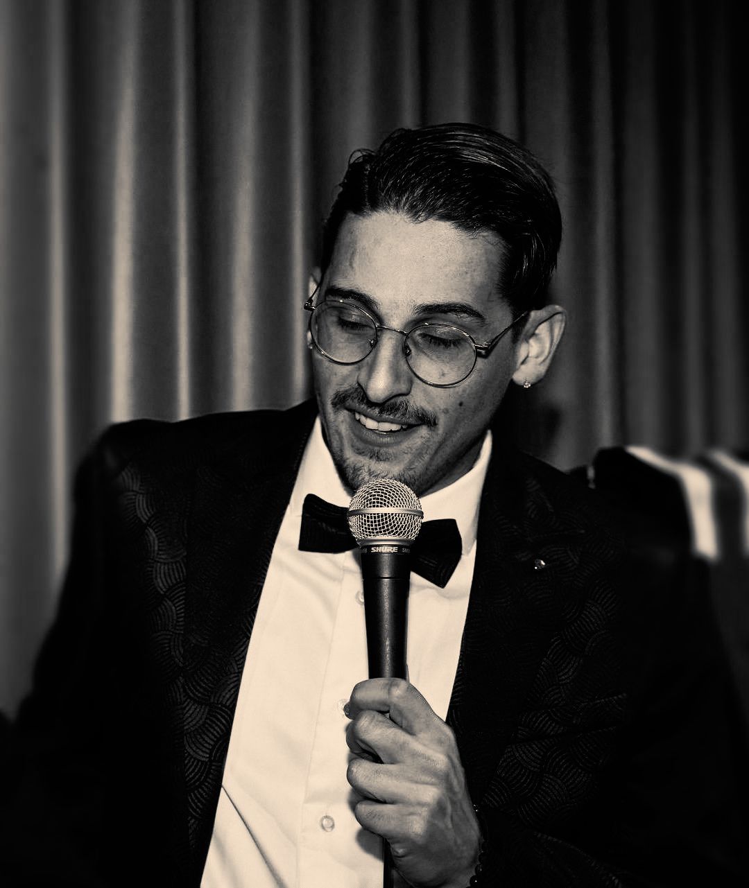 Een man in formele kleding, met een microfoon in zijn hand, lachend, spreekt waarschijnlijk op een evenement.