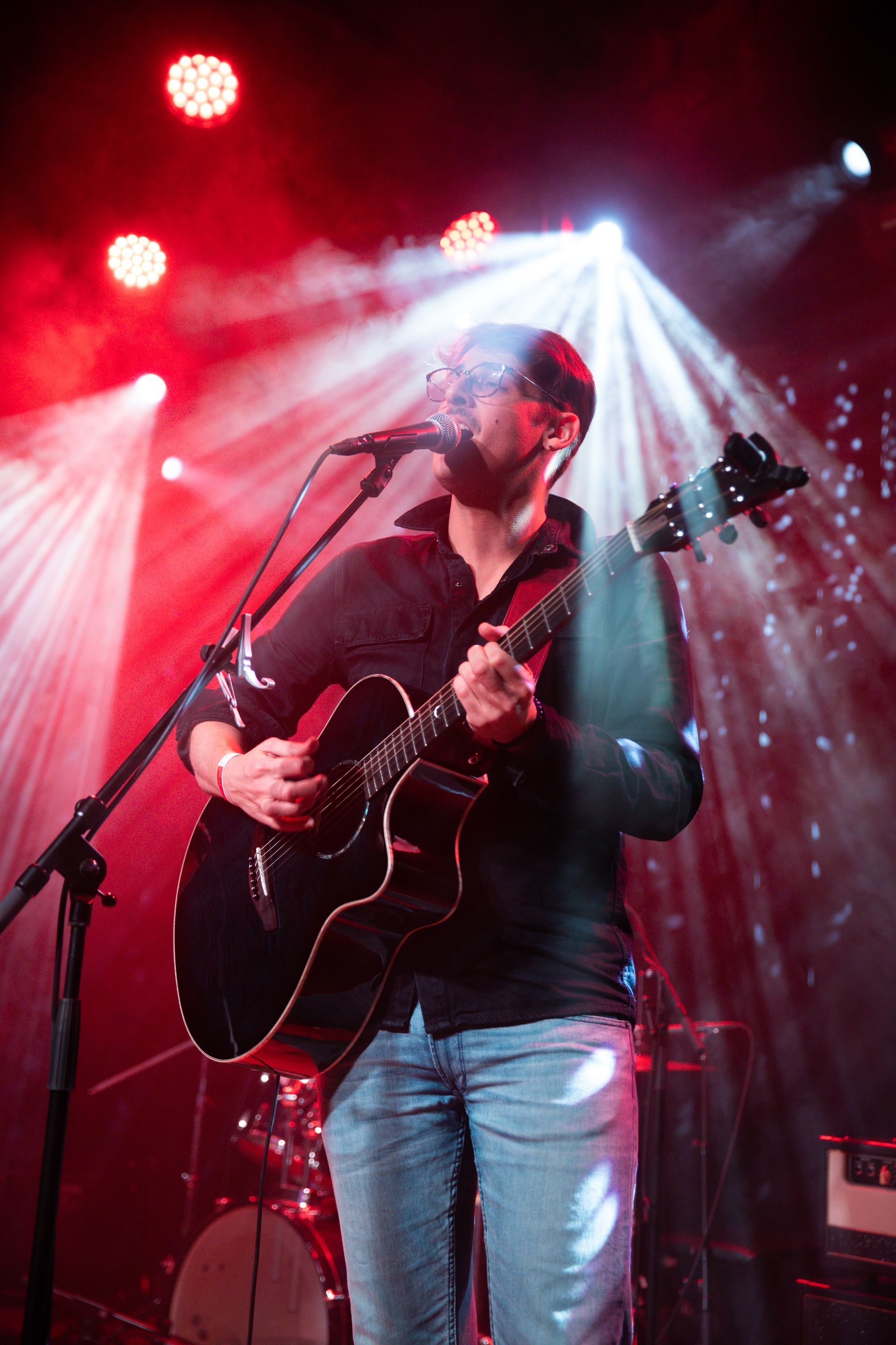 Een man speelt akoestische gitaar op het podium onder rode lichten en in rook.