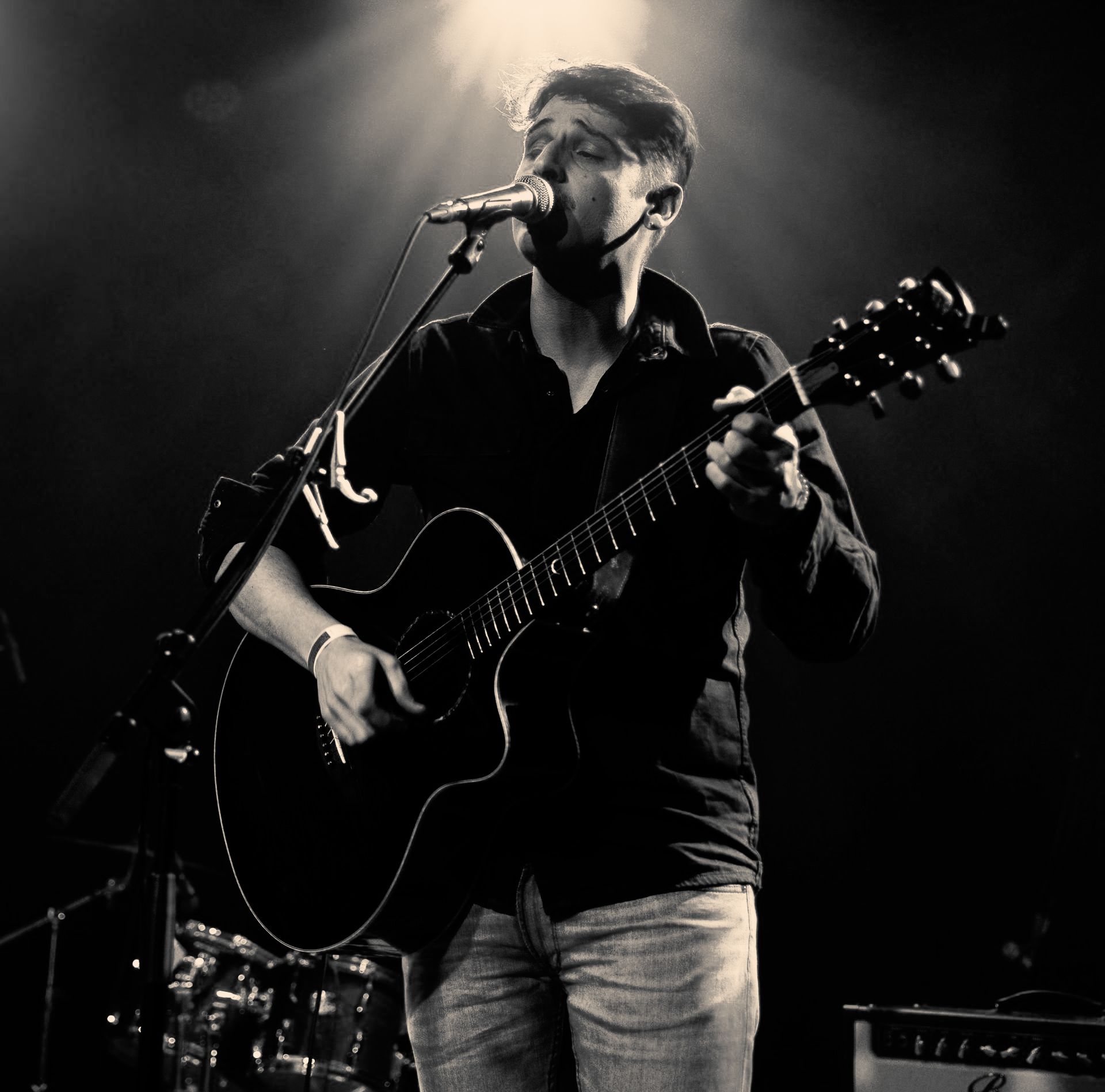 Een man zingt en speelt akoestische gitaar op het podium, onder fel podiumlicht.