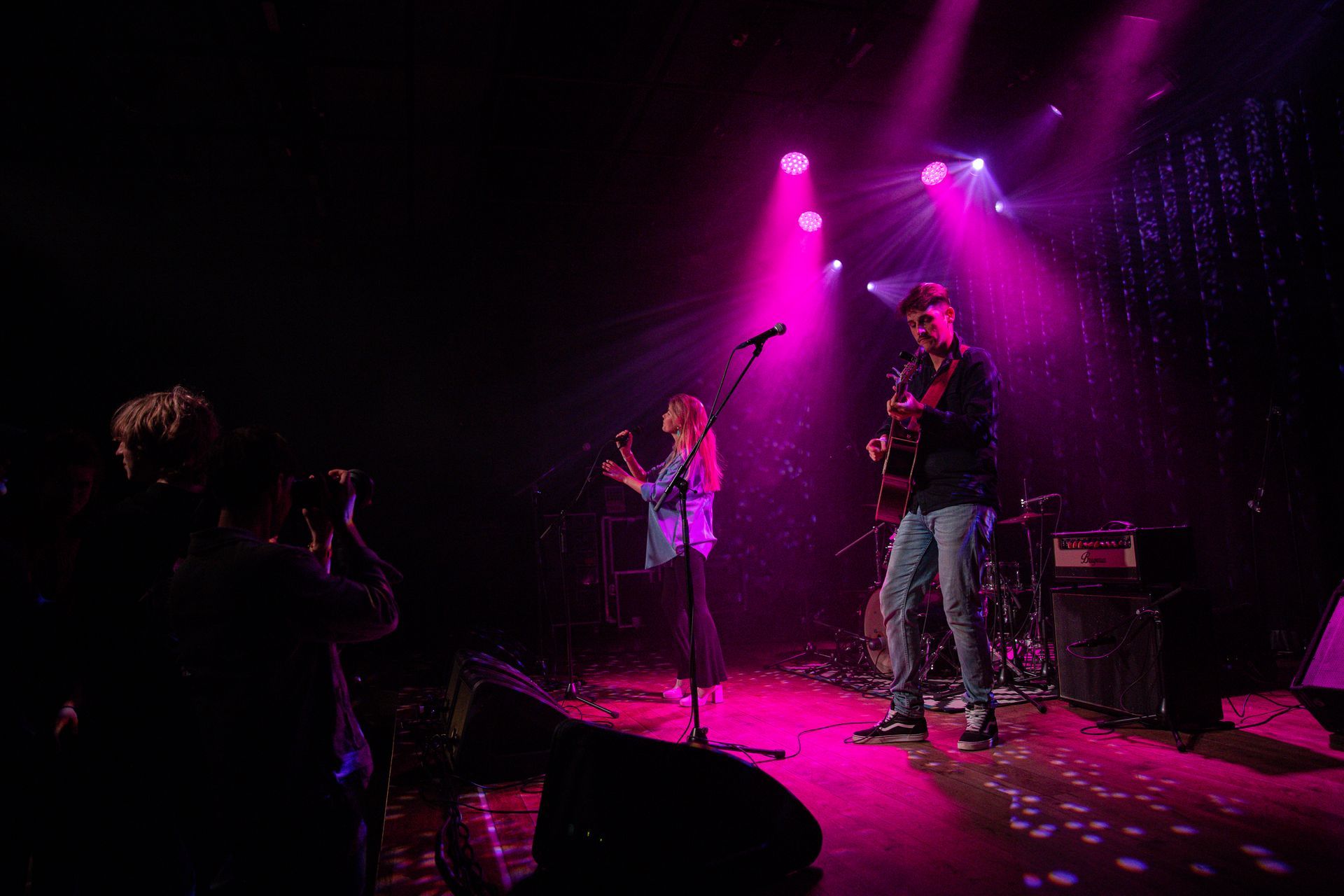 Twee muzikanten op een podium onder roze licht. Iemand aan de linkerkant maakt een foto.