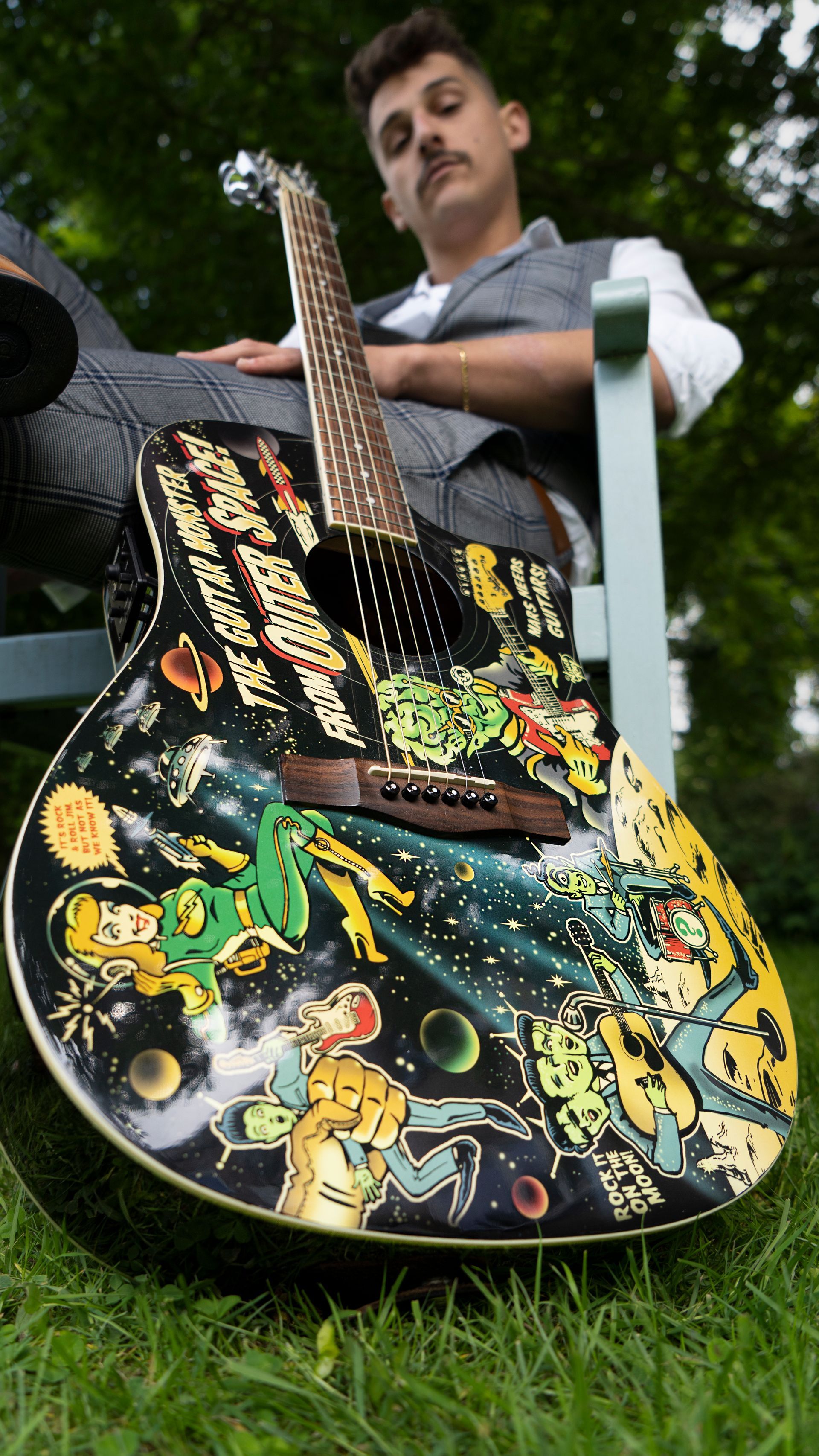 Een man zit buiten, met een kleurrijke gitaar met ruimtethema in zijn handen, en leunt tegen een stoel.