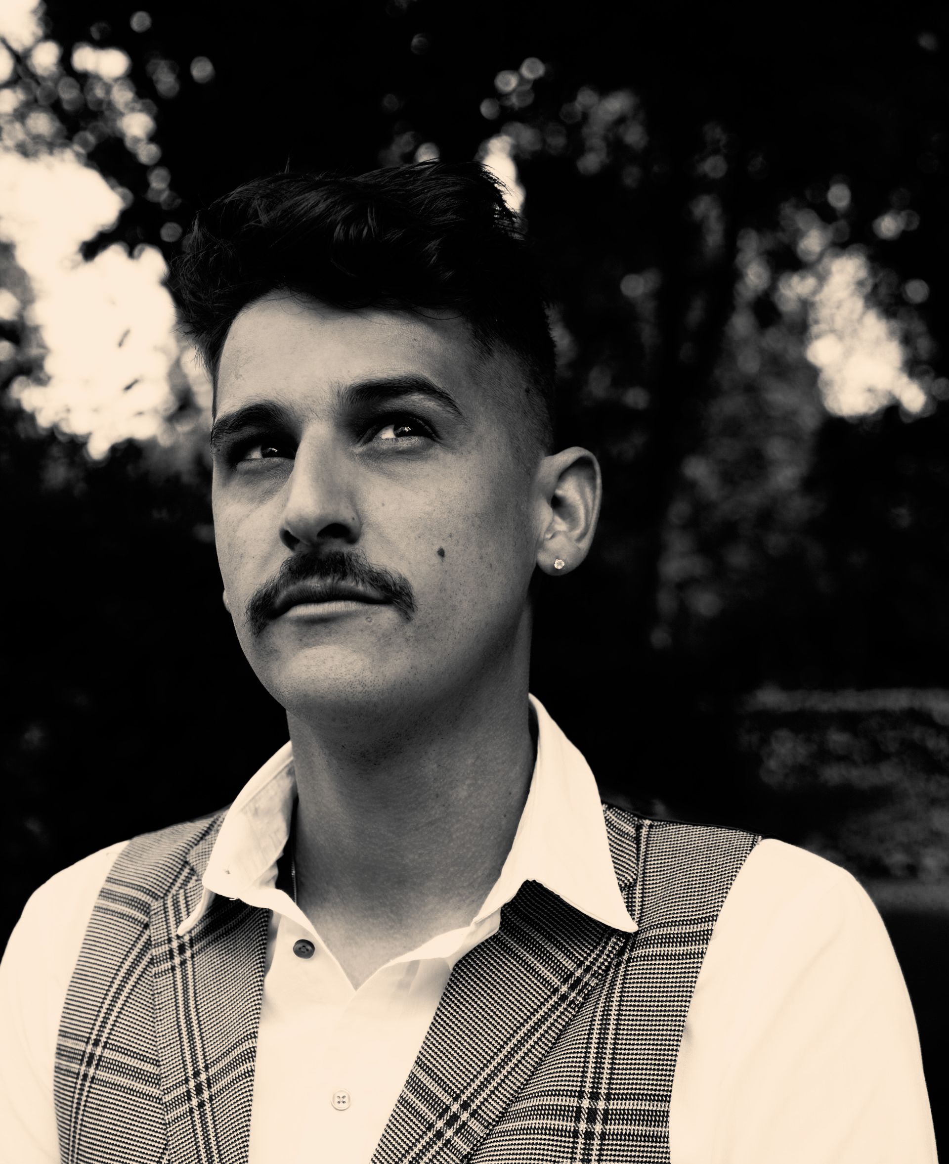 Man met snor, kijkt omhoog. Draagt ​​een geruit vest en een wit overhemd, buiten.