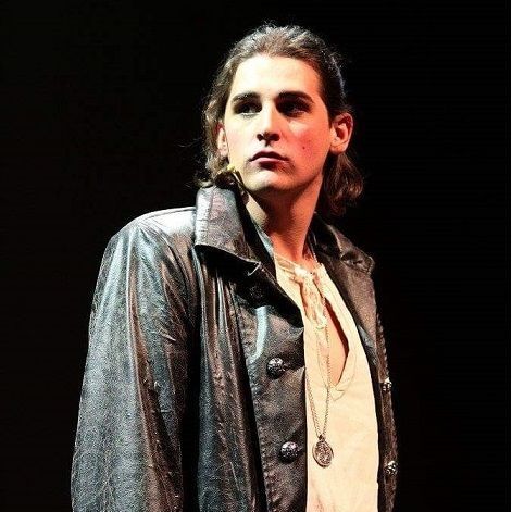 Man met lang haar, een donkere jas en een hanger, kijkt opzij. Donkere achtergrond.