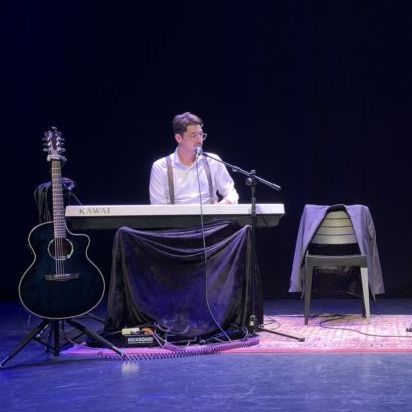Een man speelt keyboard op het podium, een gitaar ligt naast hem en zijn jas ligt op een stoel. Het podium is donker.