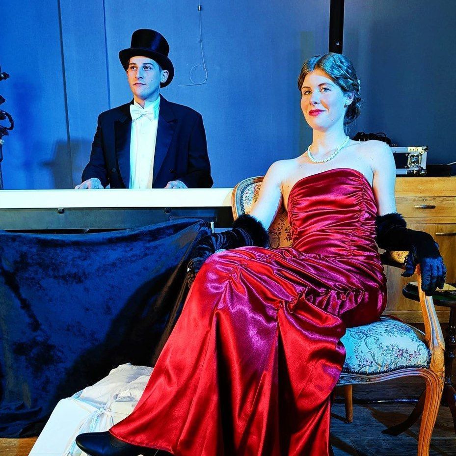 Vrouw in rode jurk zit; man in smoking op de achtergrond. Toneeldecor, schemerig verlicht.