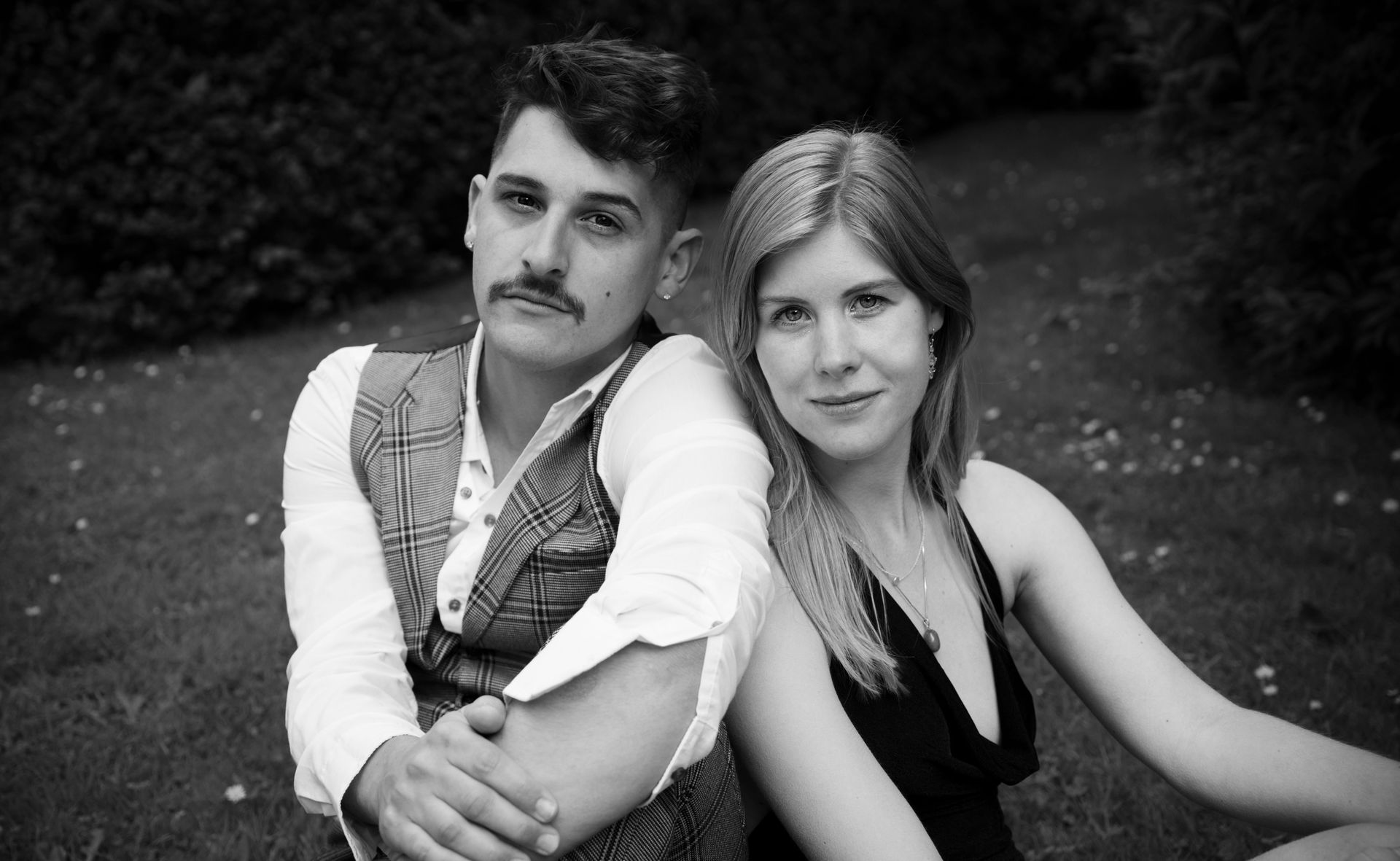 Een stel zit in het gras, de man draagt ​​een vest met snor en de vrouw een zwarte jurk; beiden glimlachen.