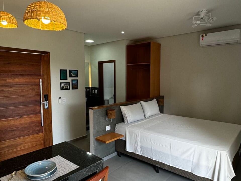 Um quarto de hotel apresenta uma cama com lençóis brancos, uma porta de madeira escura, uma pequena mesa com uma tigela e uma luminária pendente de palha trançada.