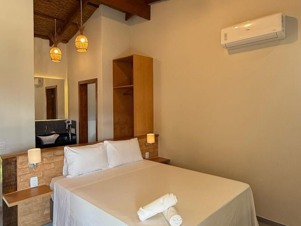 Um quarto de hotel com cama de lençóis brancos, detalhes em madeira, luminárias pendentes, espelho e ar-condicionado de parede.