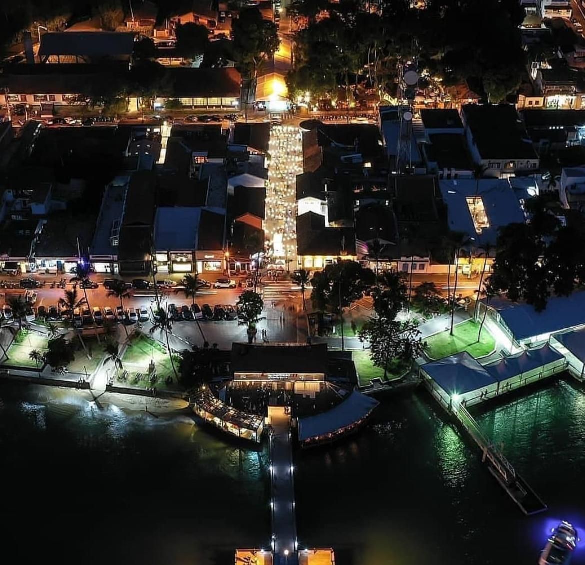 Vista aérea noturna de uma cidade litorânea bem iluminada, com um píer que se estende pelas águas escuras, cercada por árvores exuberantes.